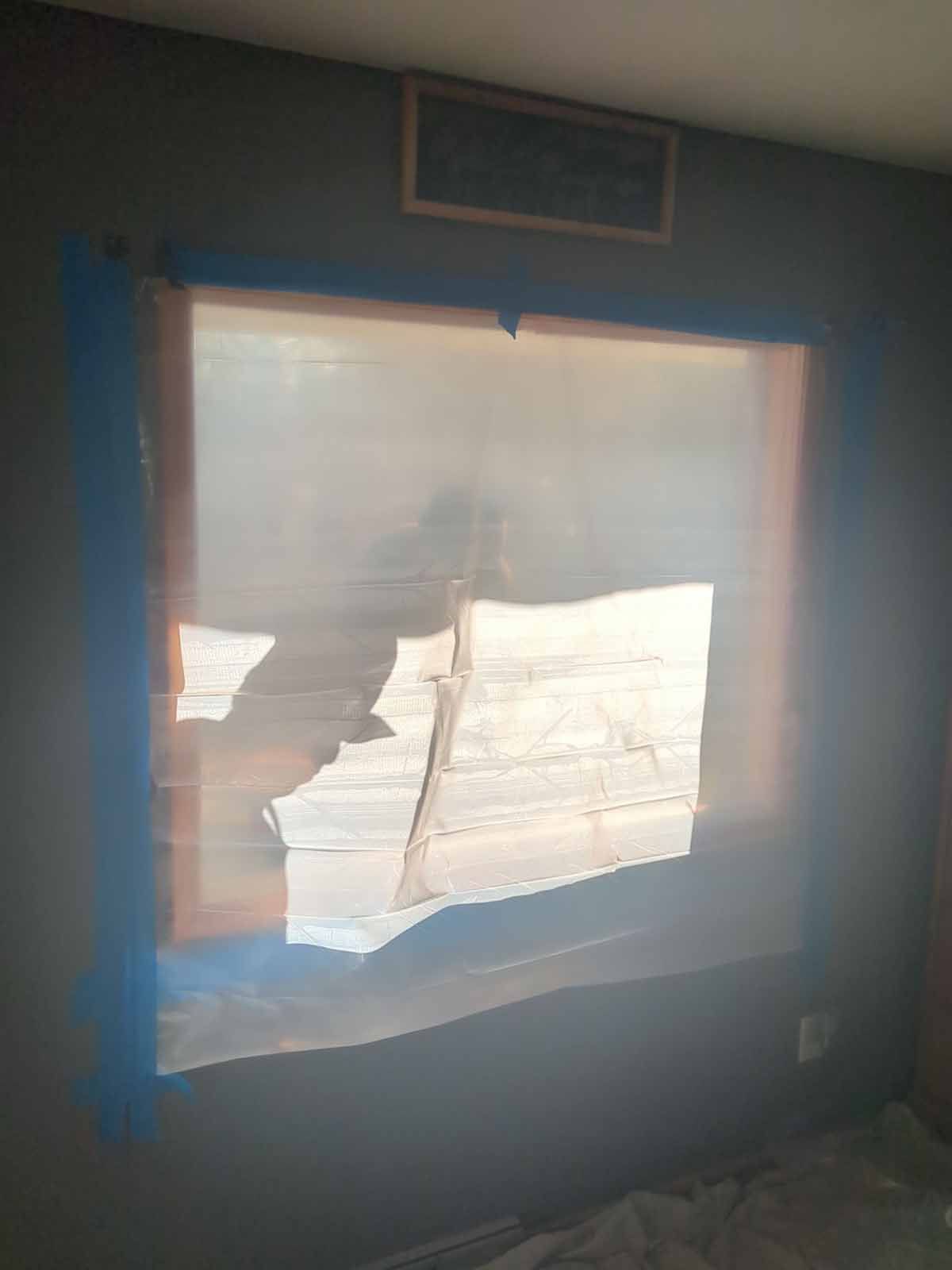 Window covered in plastic sheeting, blue tape along edges. Sunlight streams through.
