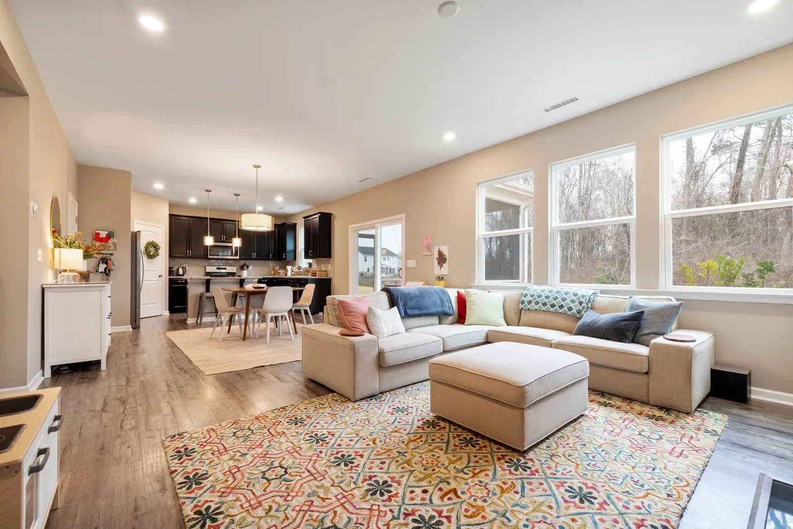 Open-concept living room with neutral colors, a colorful rug, and large windows overlooking trees.