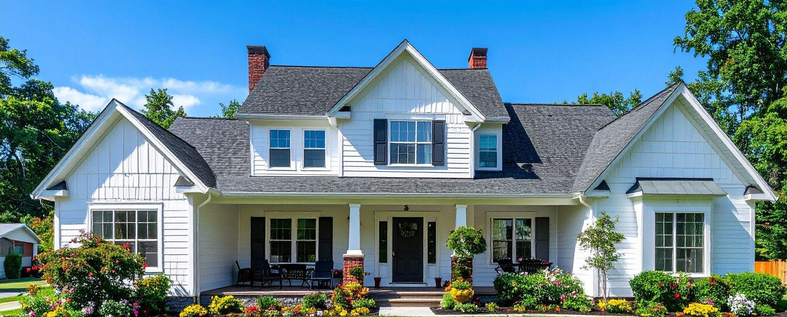 White farmhouse with black shutters, gray roof, and a porch on a sunny day.