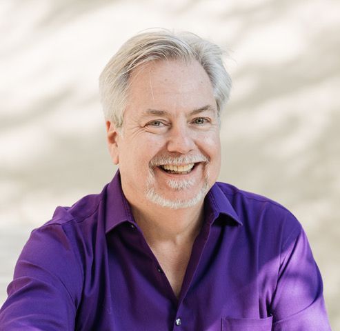 A man with a beard is wearing a purple shirt and smiling.