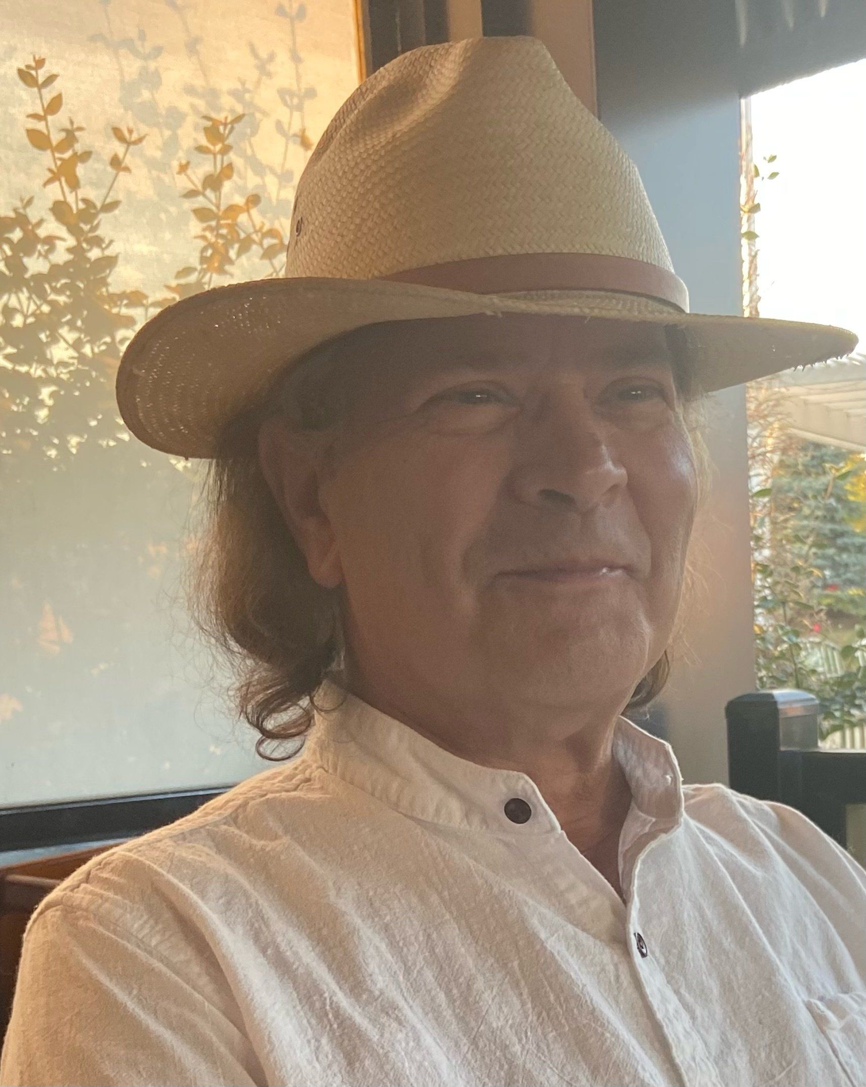 A man wearing a hat and a white shirt is sitting in front of a window.