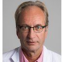 A man wearing glasses and a white coat is looking at the camera.