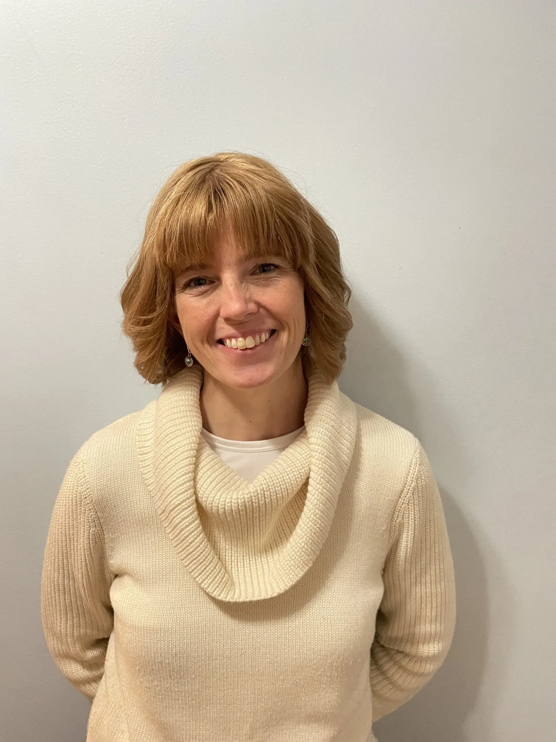 A woman in a white sweater is smiling and standing in front of a white wall.