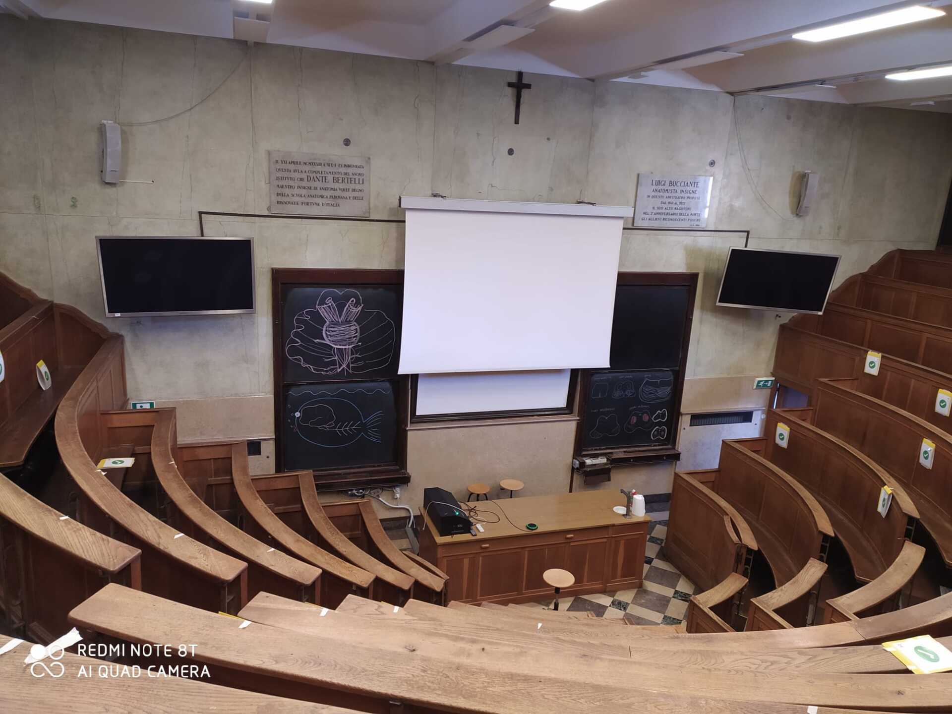 An empty lecture hall with rows of wooden seats and a projector screen.