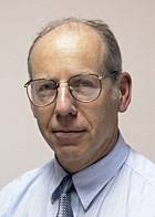 A man wearing glasses and a blue shirt and tie is looking at the camera.