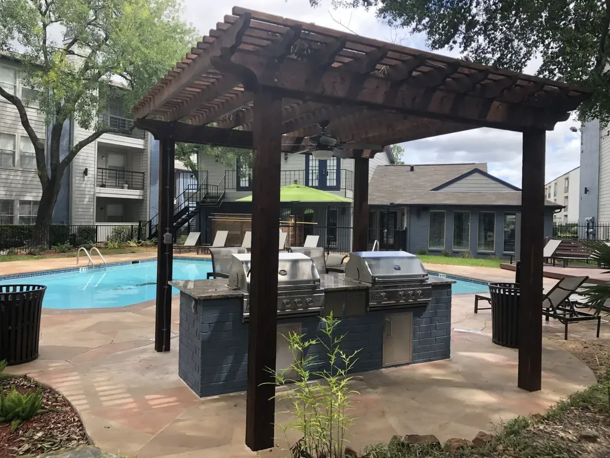 There is a grill under a pergola next to a pool.