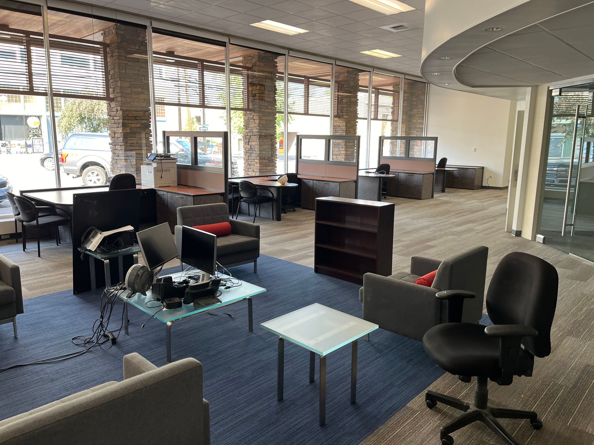 Modern office lounge with blue carpet, chairs, tables, and large windows with daylight.