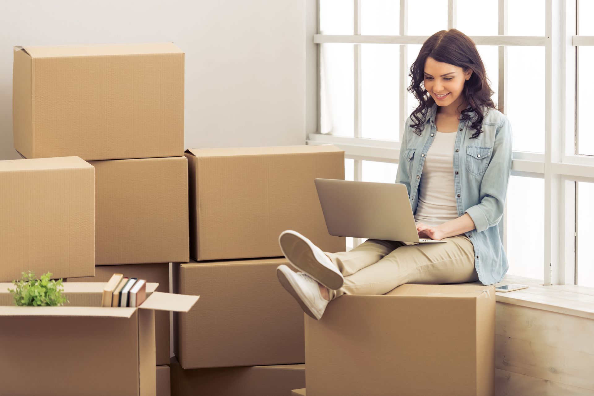 Woman using a laptop, sitting on moving boxes near a window; smiling, relaxed.