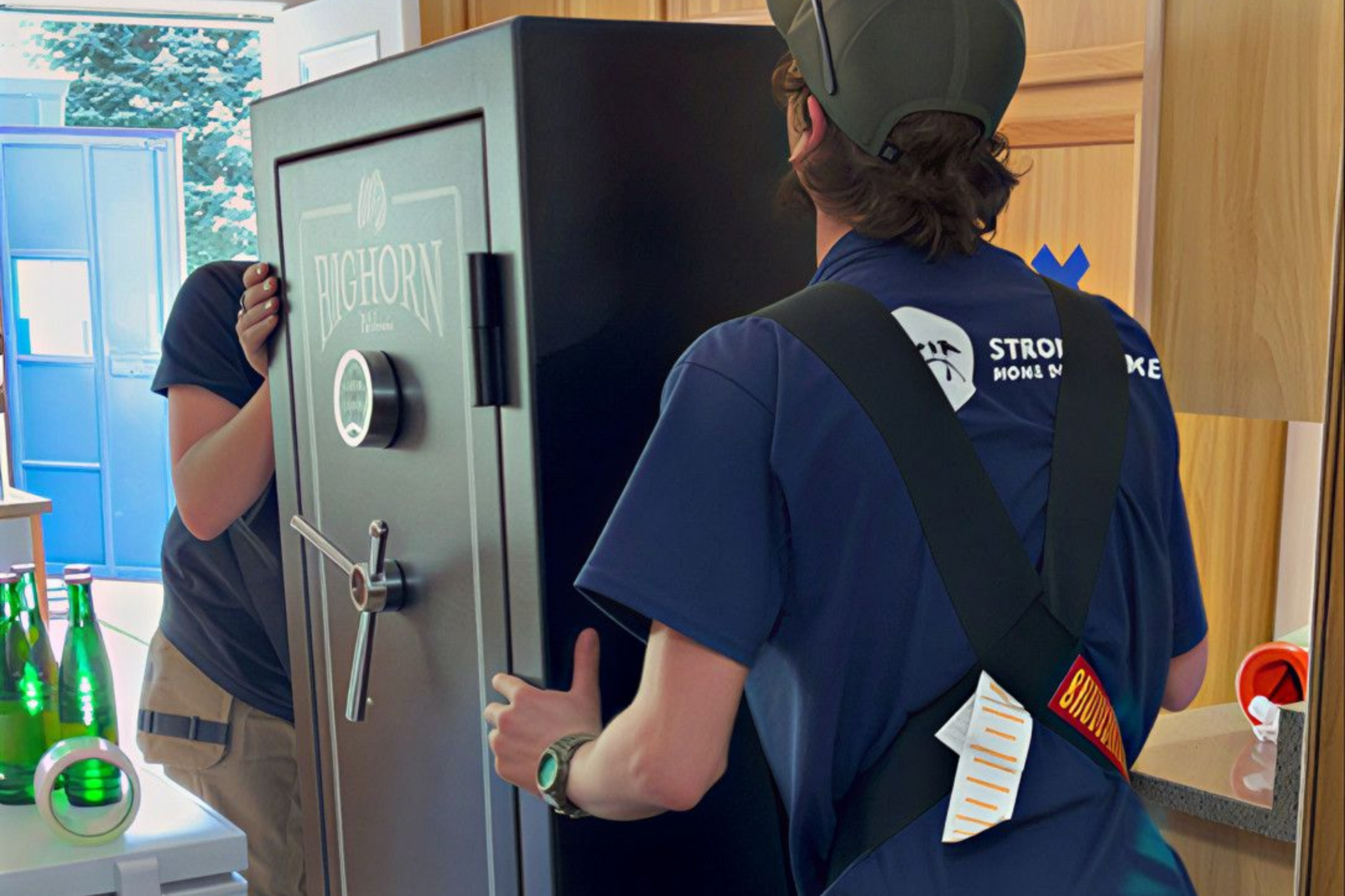 Two movers carrying a large black safe through a doorway in a kitchen.
