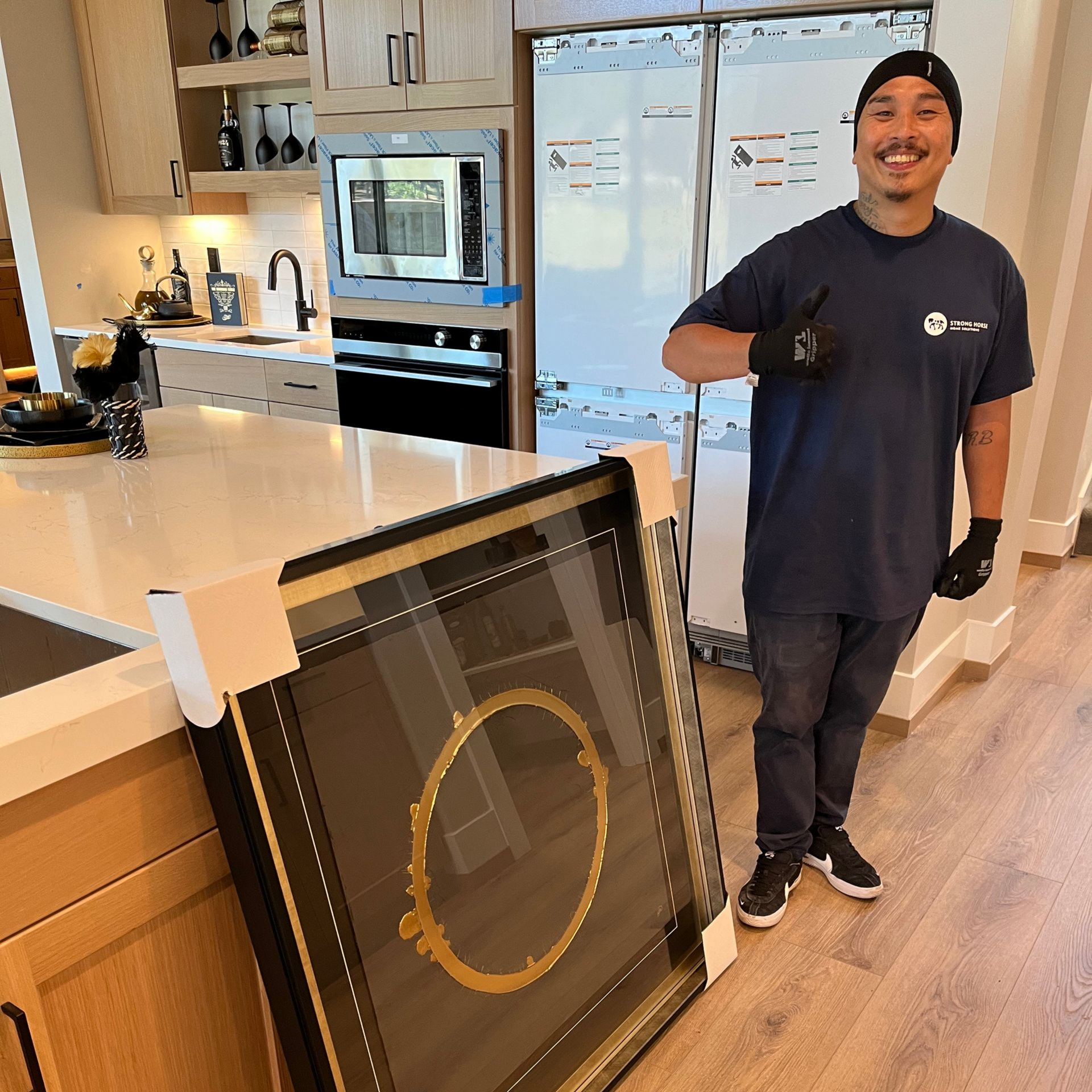 Smiling person holds packaged artwork in a kitchen, giving a thumbs up.
