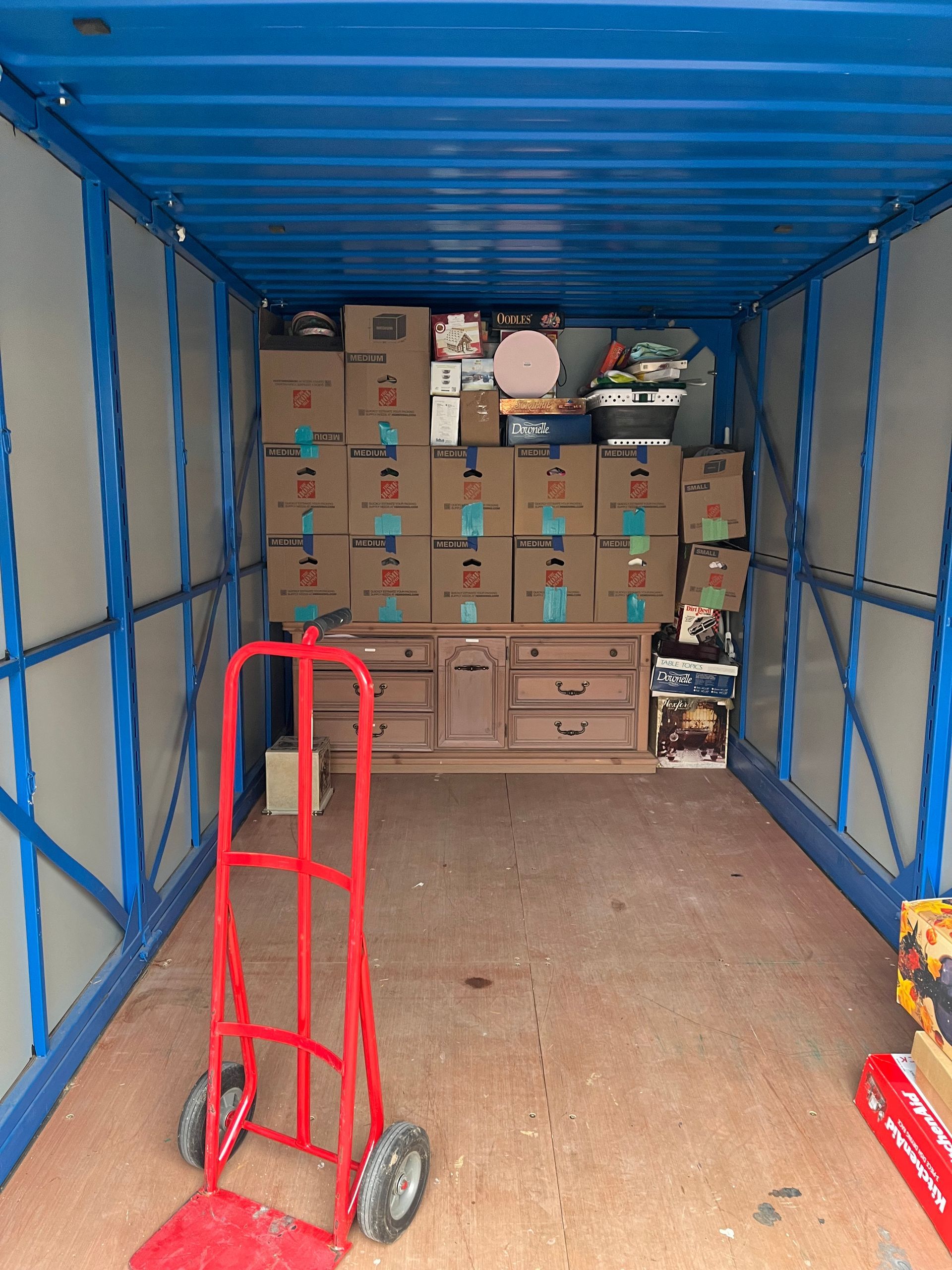 Interior of a storage container, filled with boxes and furniture. A red hand truck is in the foreground.
