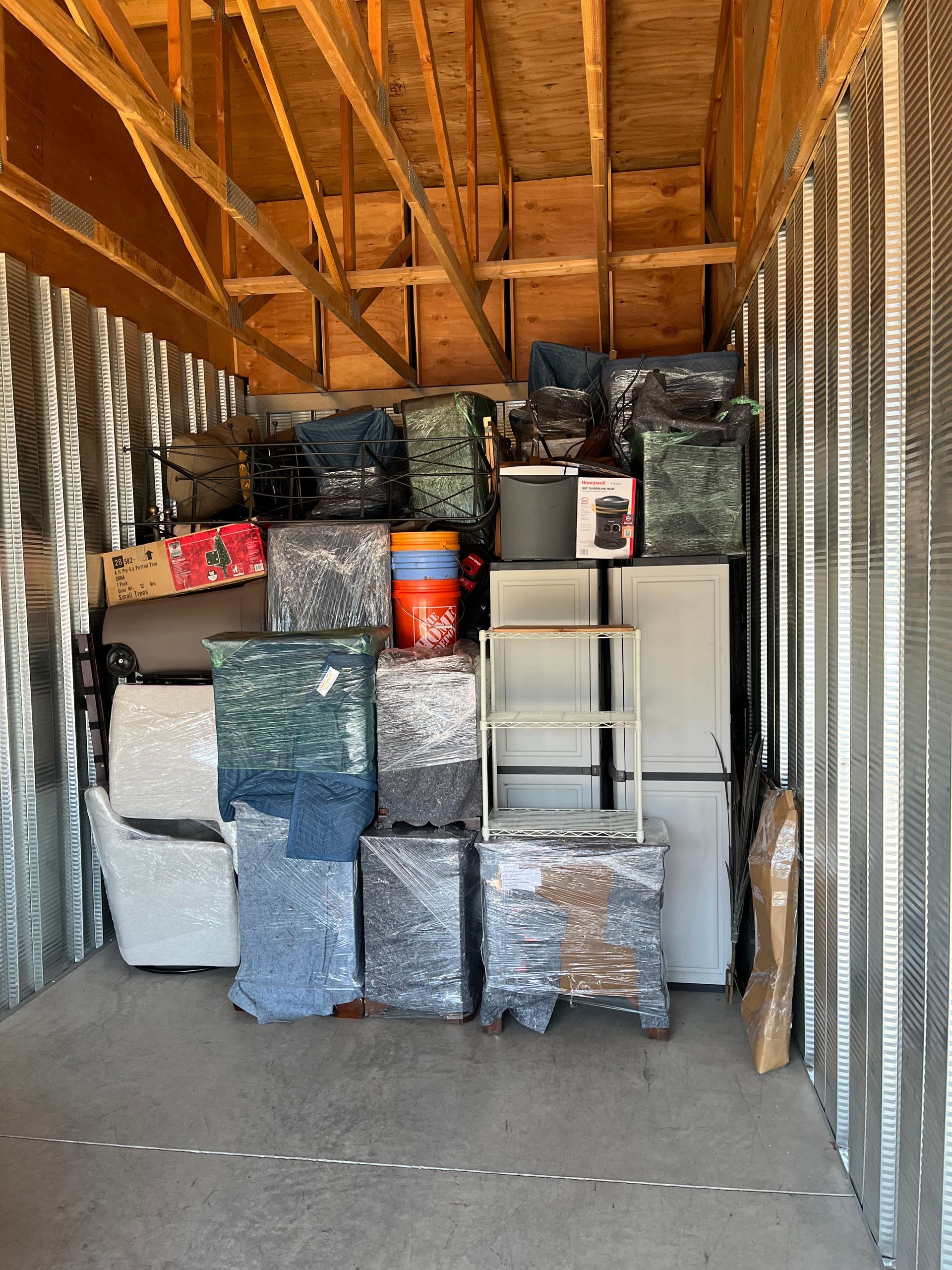 Storage unit packed with furniture and boxes, some wrapped in plastic. Wooden ceiling, corrugated metal walls.