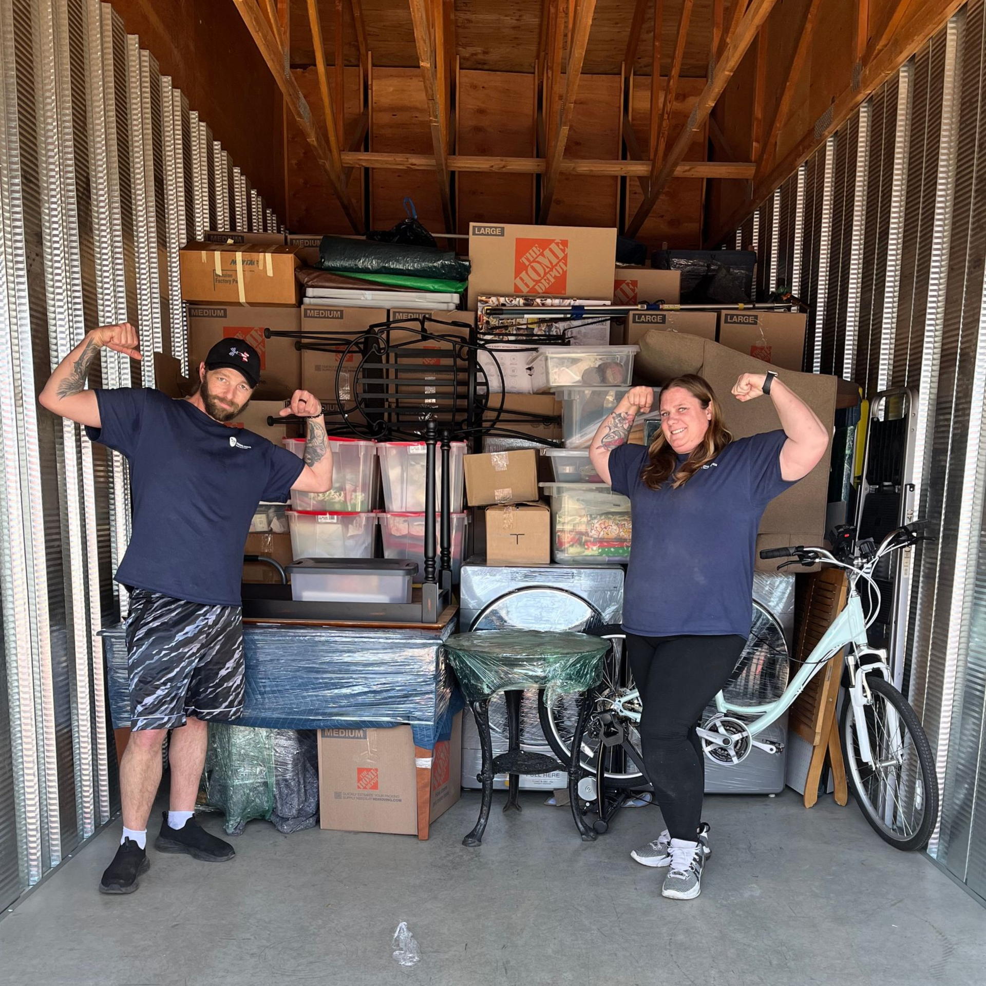Two people flexing muscles in a packed storage unit. Boxes, furniture, and a bike are visible.
