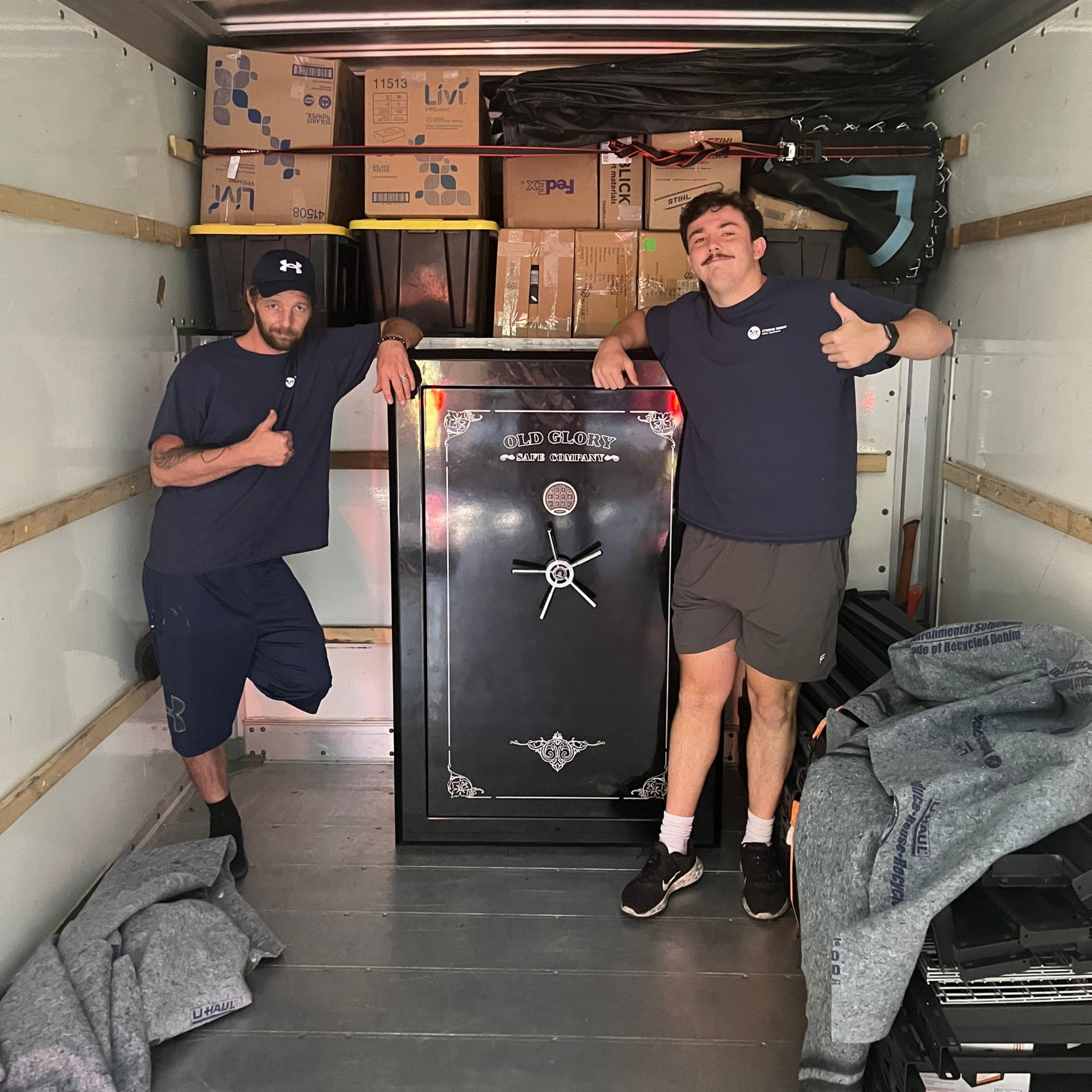 Two men in a moving truck with a large safe and boxes, both giving thumbs up.