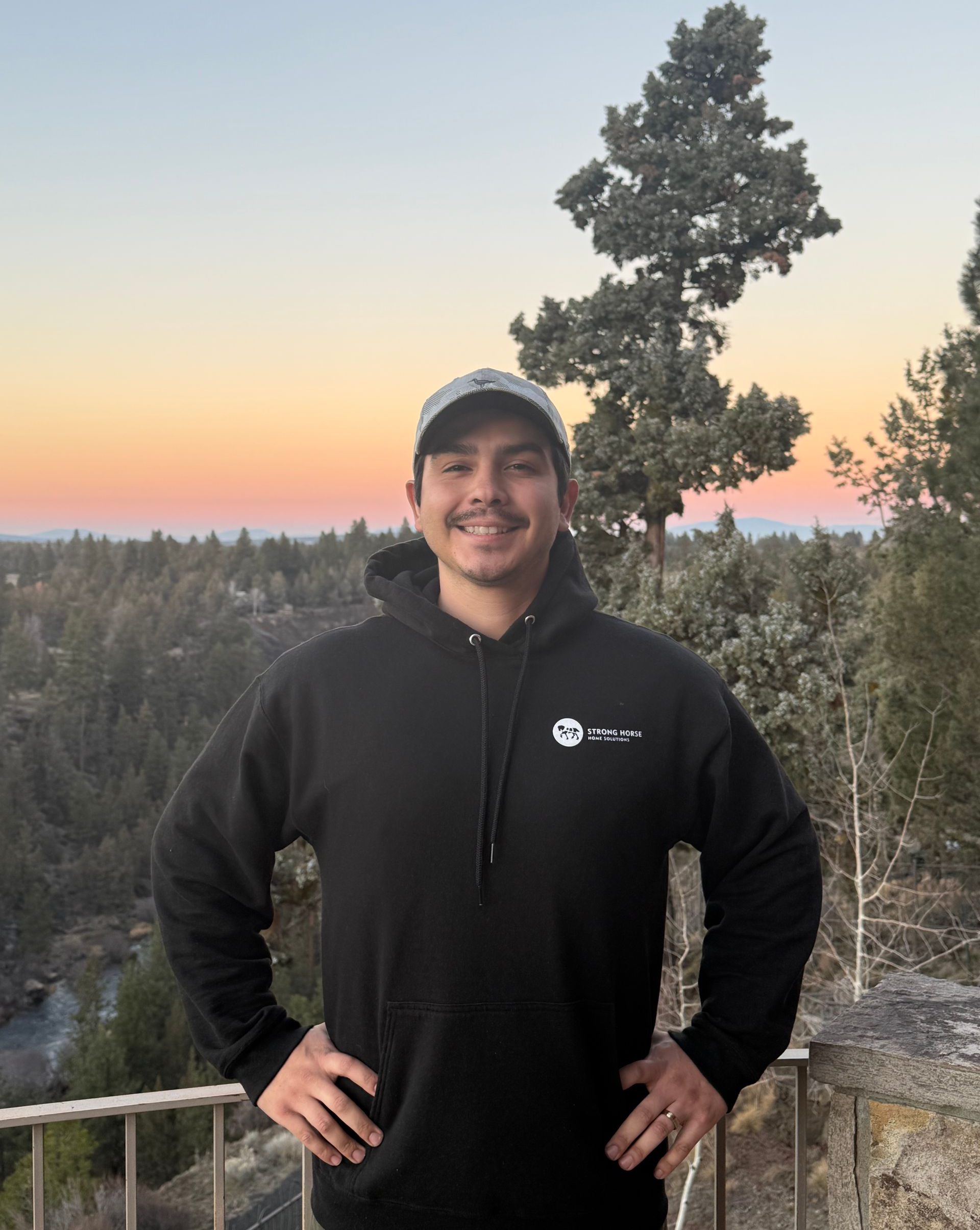 Man in black hoodie with hands on hips, standing outdoors with a sunset backdrop.