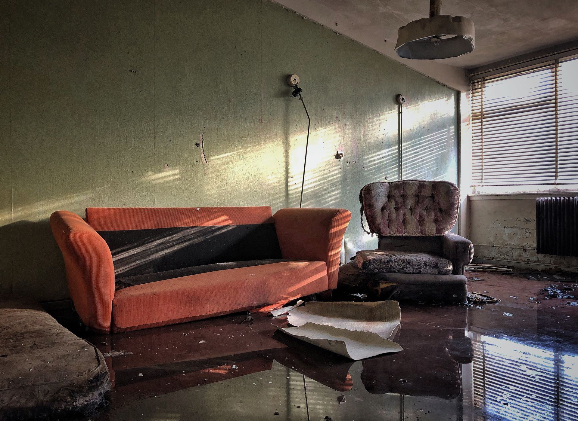 Abandoned, dilapidated living room with orange couch, armchair, and flooded floor; sunlight through blinds.