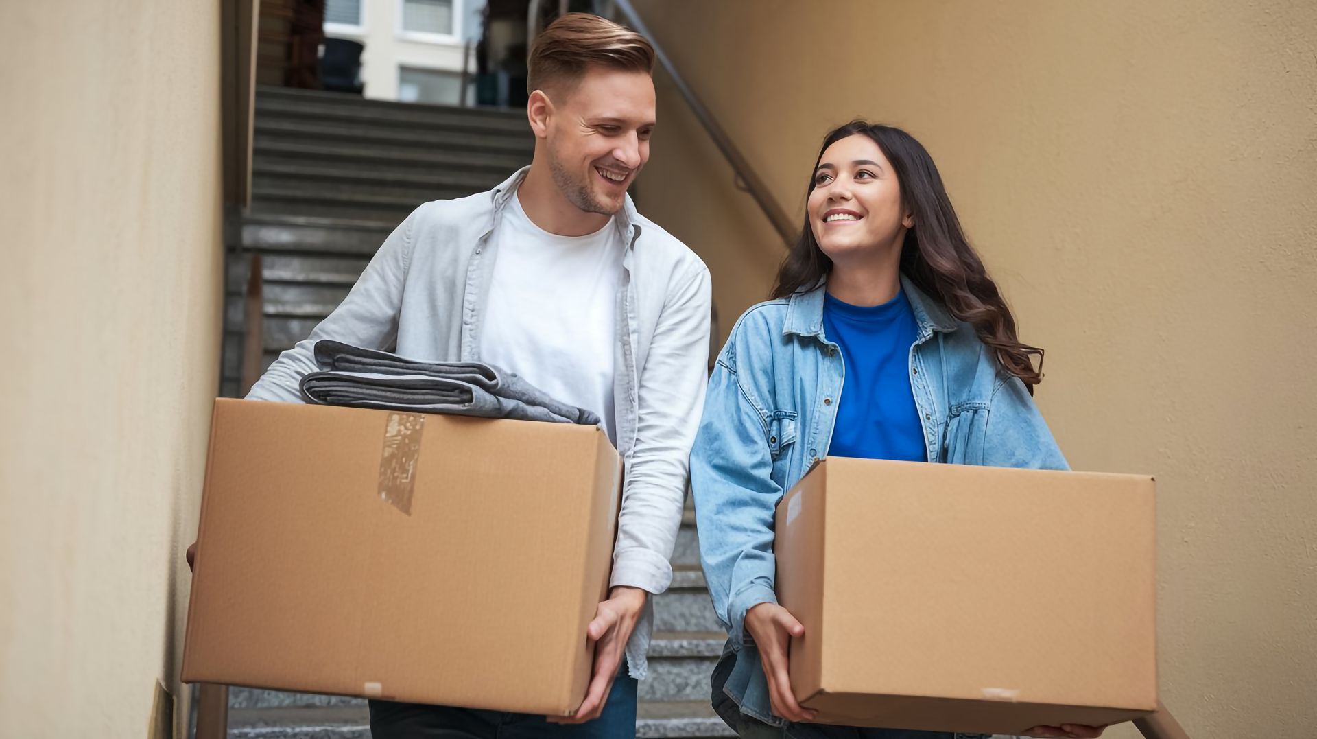 Couple moving boxes down stairs, smiling at each other.
