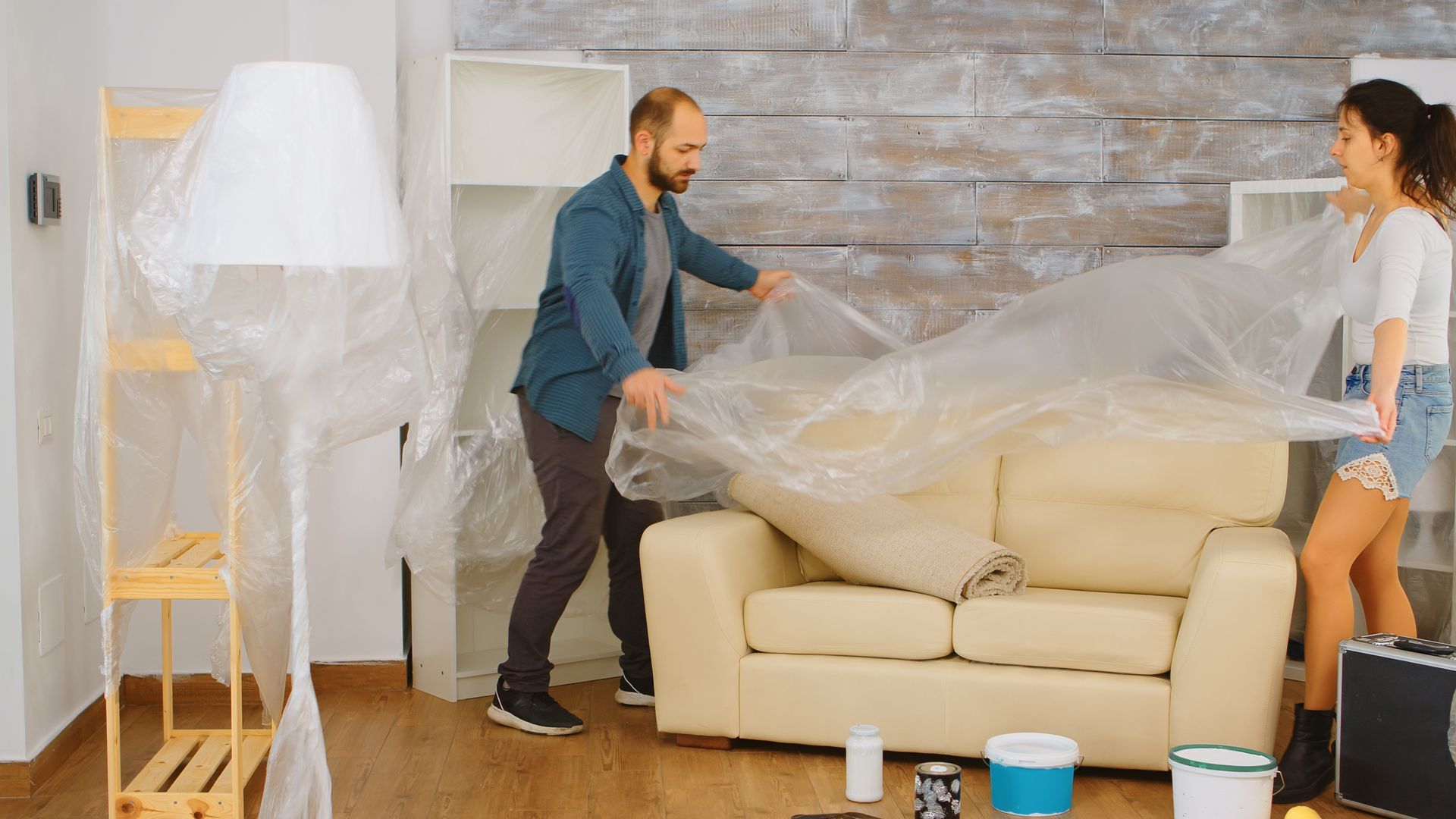 Couple wrapping couch in plastic during a move. Light-colored sofa, light wood floor, bright room.