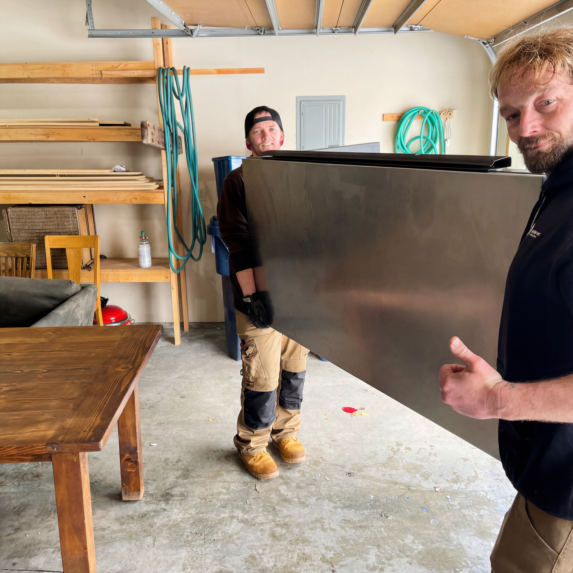 Two men carrying a large, silver metal object in a garage, one giving a thumbs up.