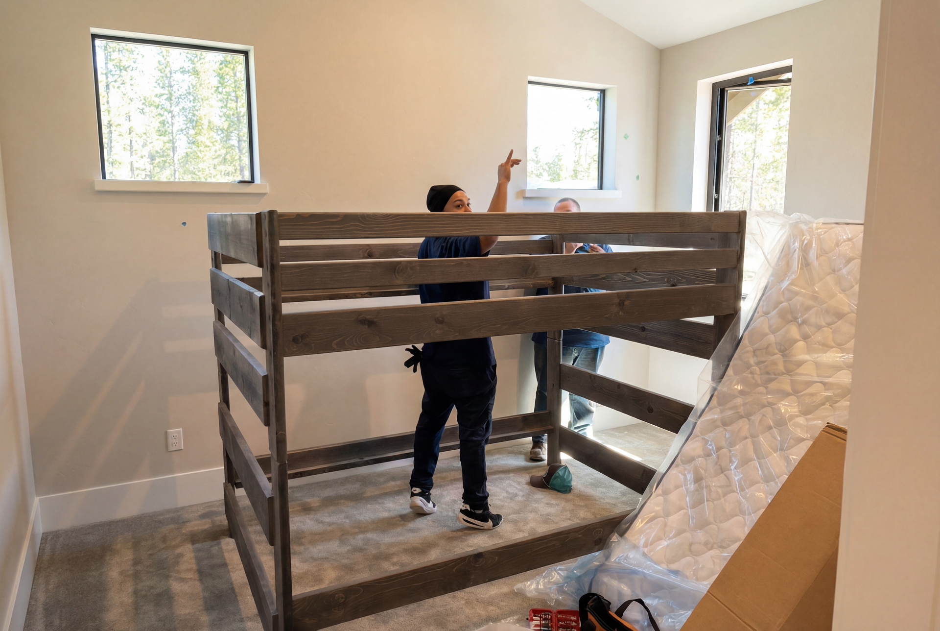 Two people assemble a gray wooden bunk bed in a bedroom with natural light.