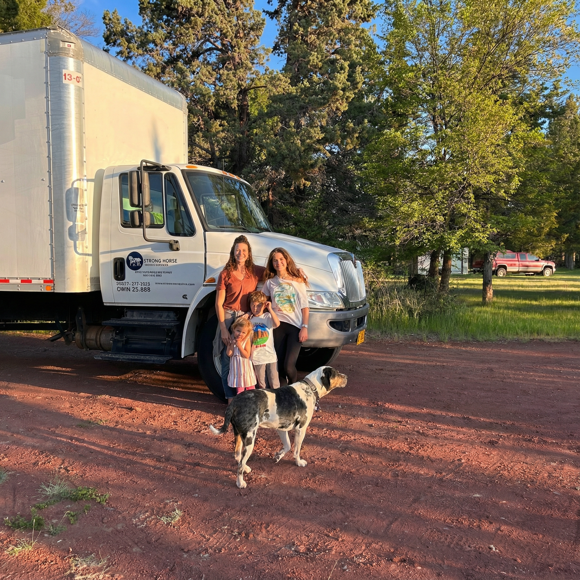 Family with a dog in front of a moving truck on a dirt road, trees in the background.