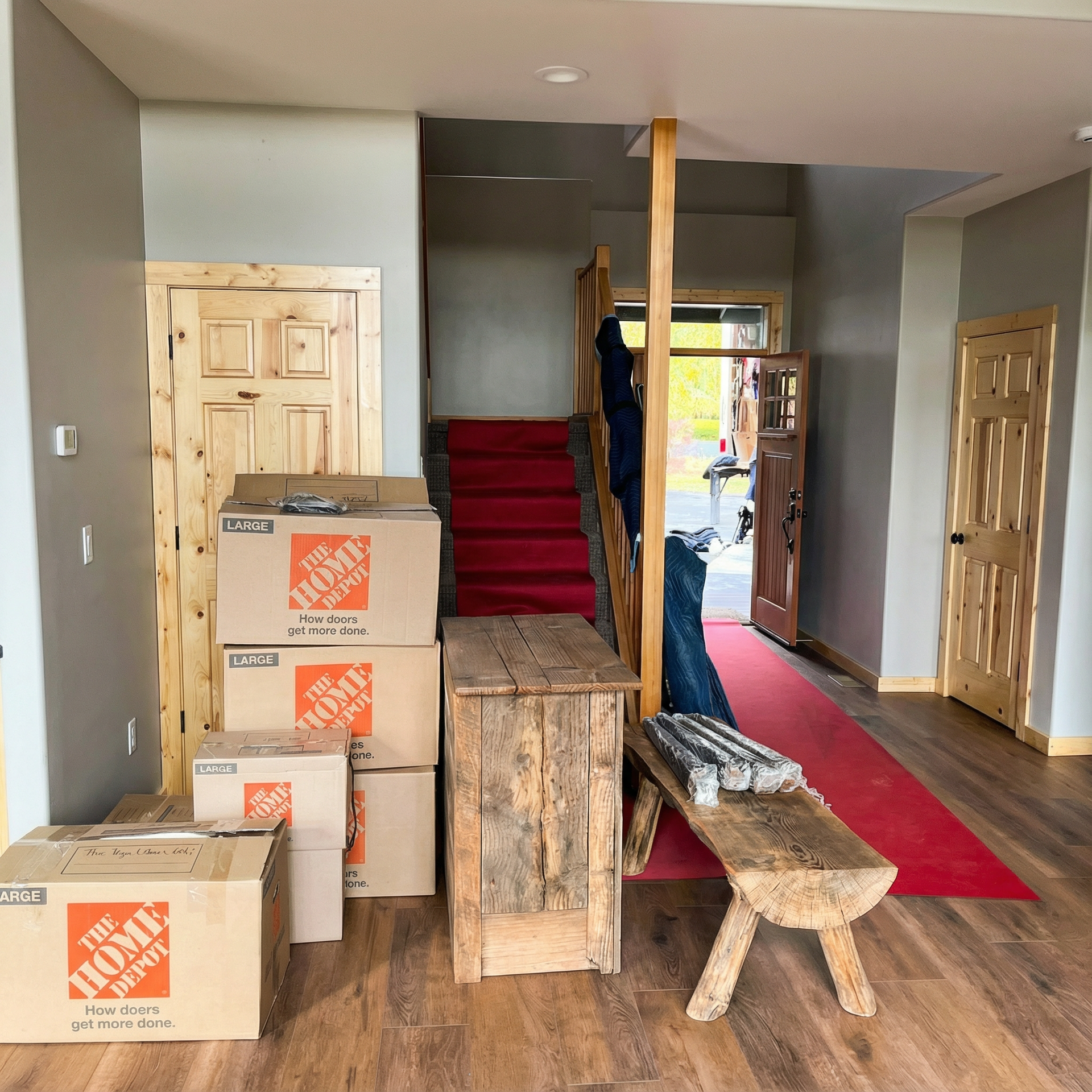 Entryway with moving boxes, a rustic bench, stairs with red carpet, and open doors.