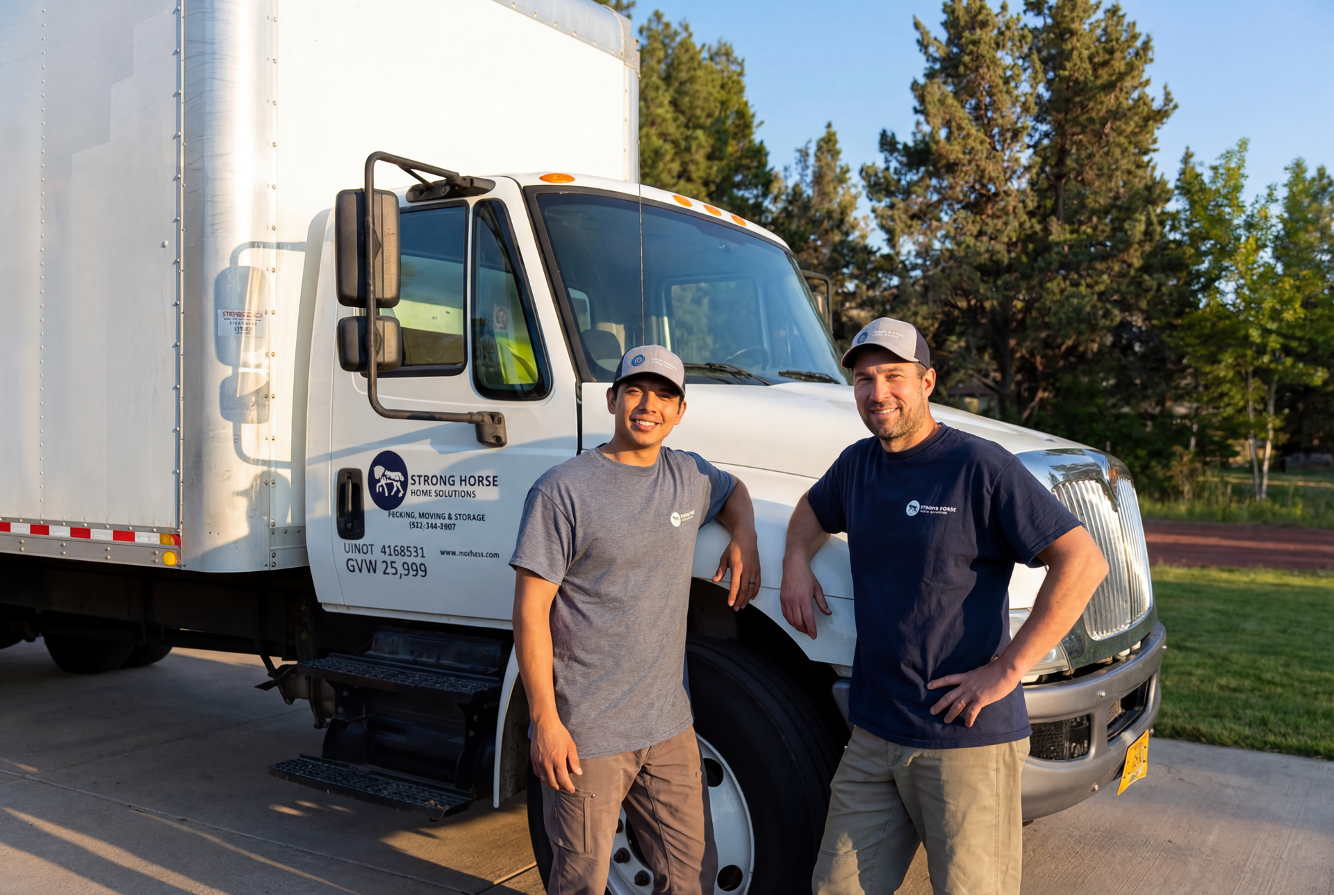 Two movers stand beside a white moving truck. One leans on the truck, the other has hands on hips, both wearing matching shirts.