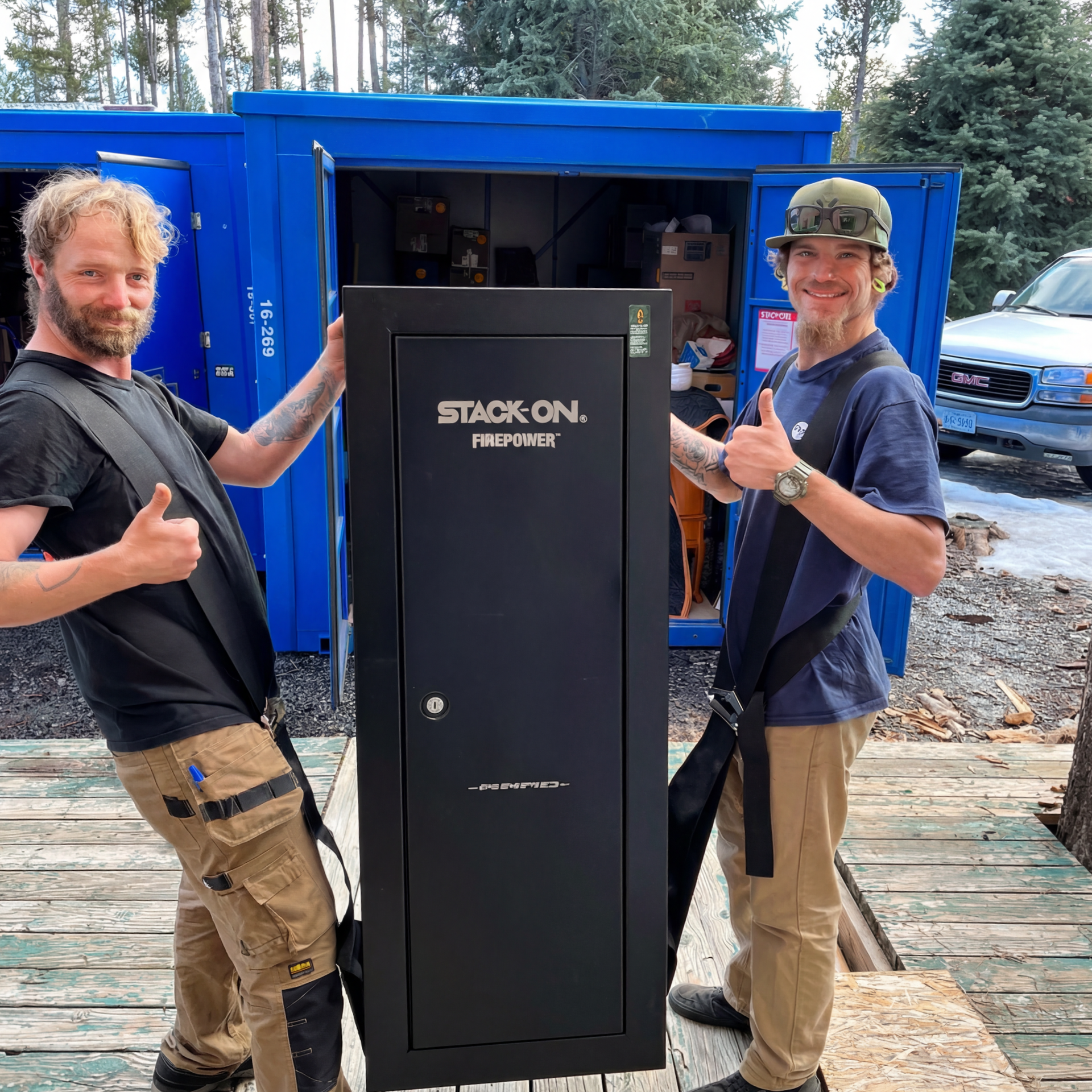 Two men holding a large black Stack-On safe, thumbs up, near blue containers and a truck.