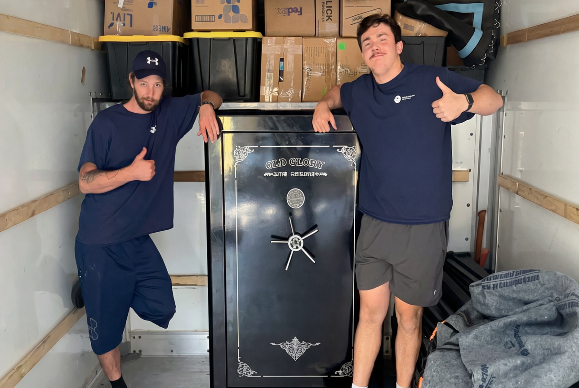 Two men in a moving truck, smiling and giving thumbs-up next to a large black safe.