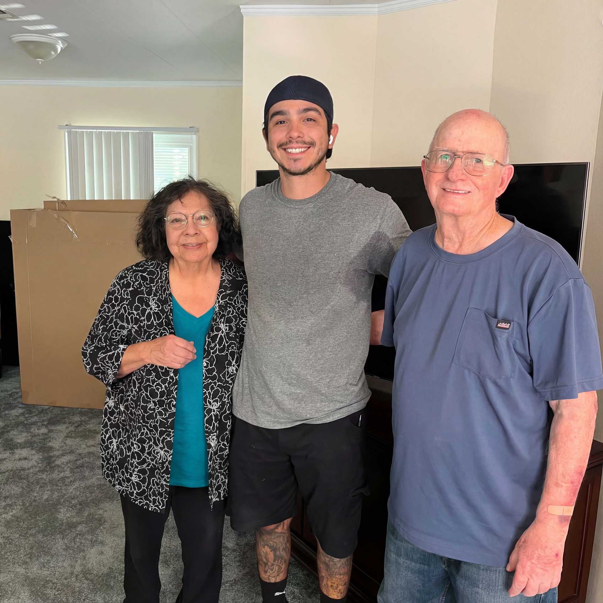Three people pose for a photo in a living room. A man with a hat, an elderly woman, and an elderly man smile.