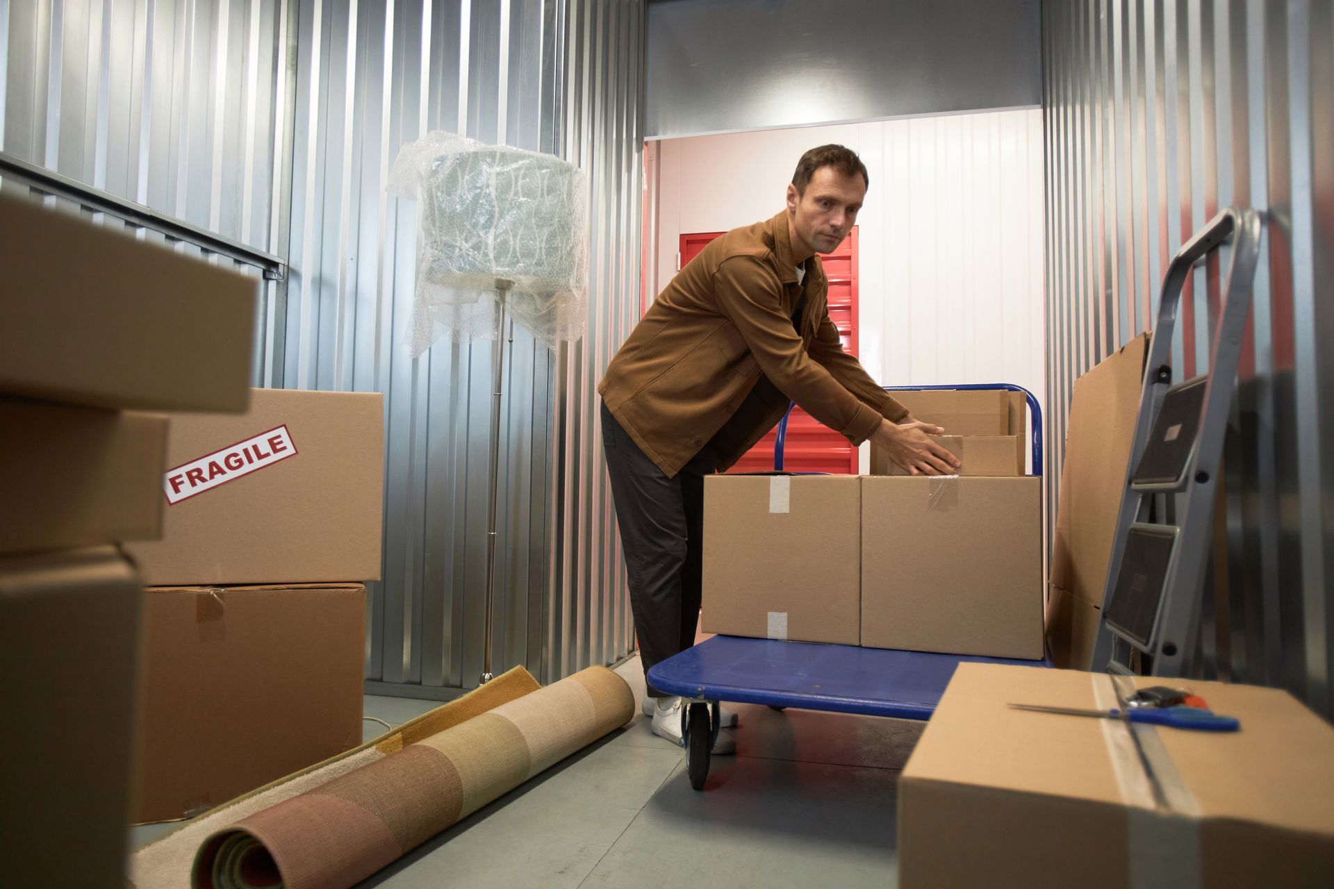 Man in a storage unit loading boxes onto a cart. Brown shirt, cardboard boxes, metal walls, and a rolled rug are visible.