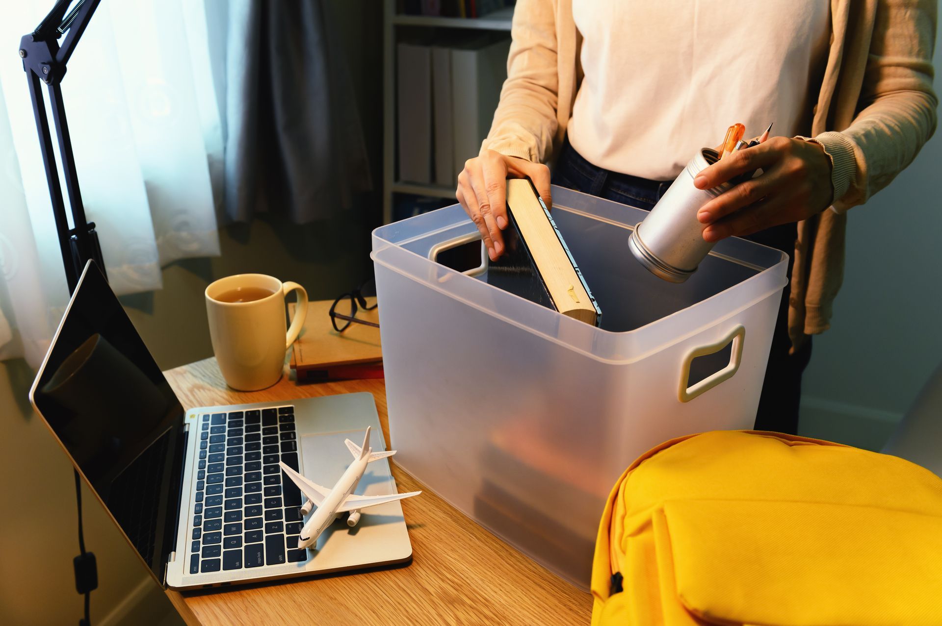A woman is standing in front of a desk with a laptop and a plastic bin.