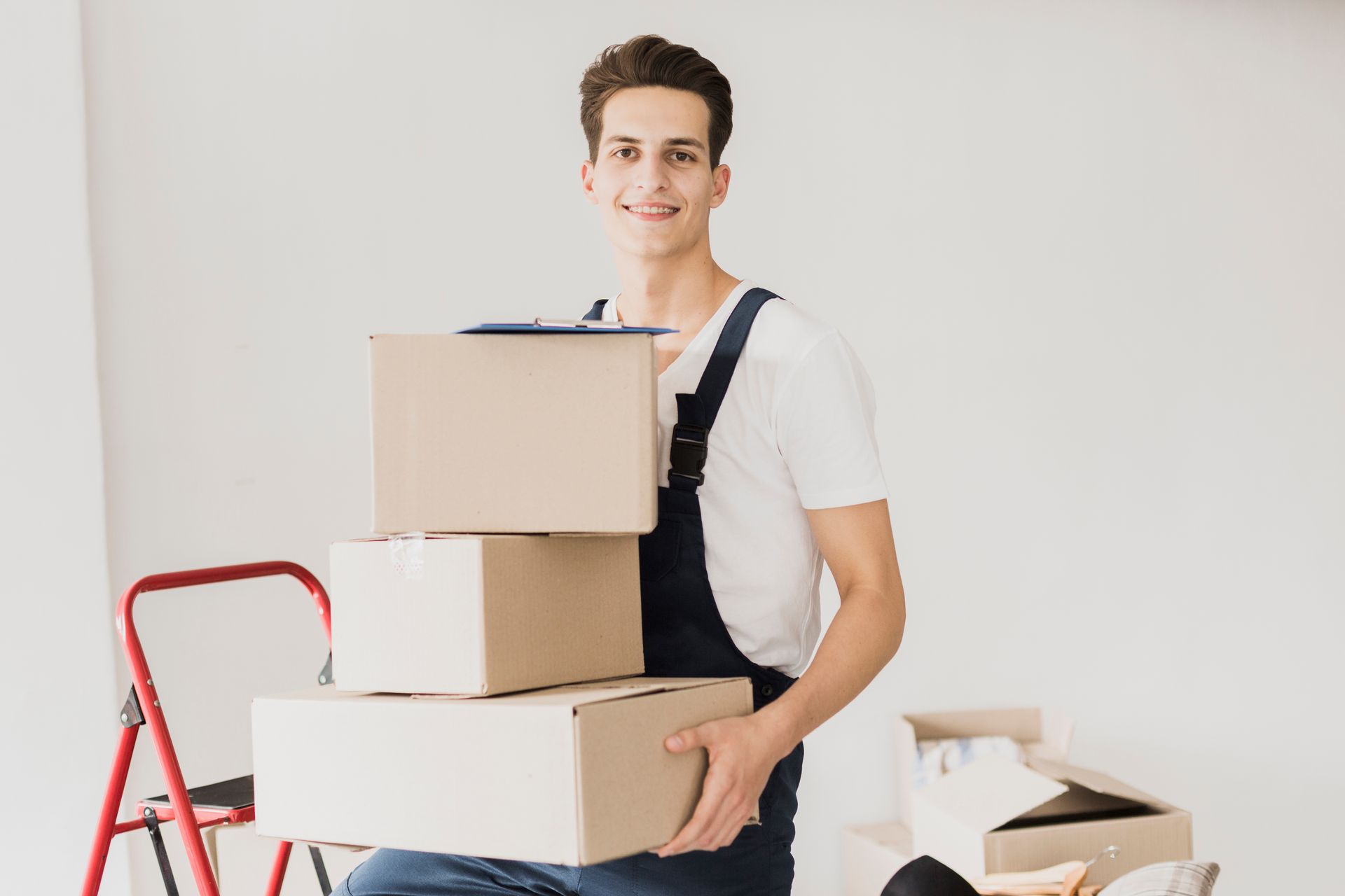 Man in overalls smiles, holding stacked cardboard boxes. White interior, red ladder visible.