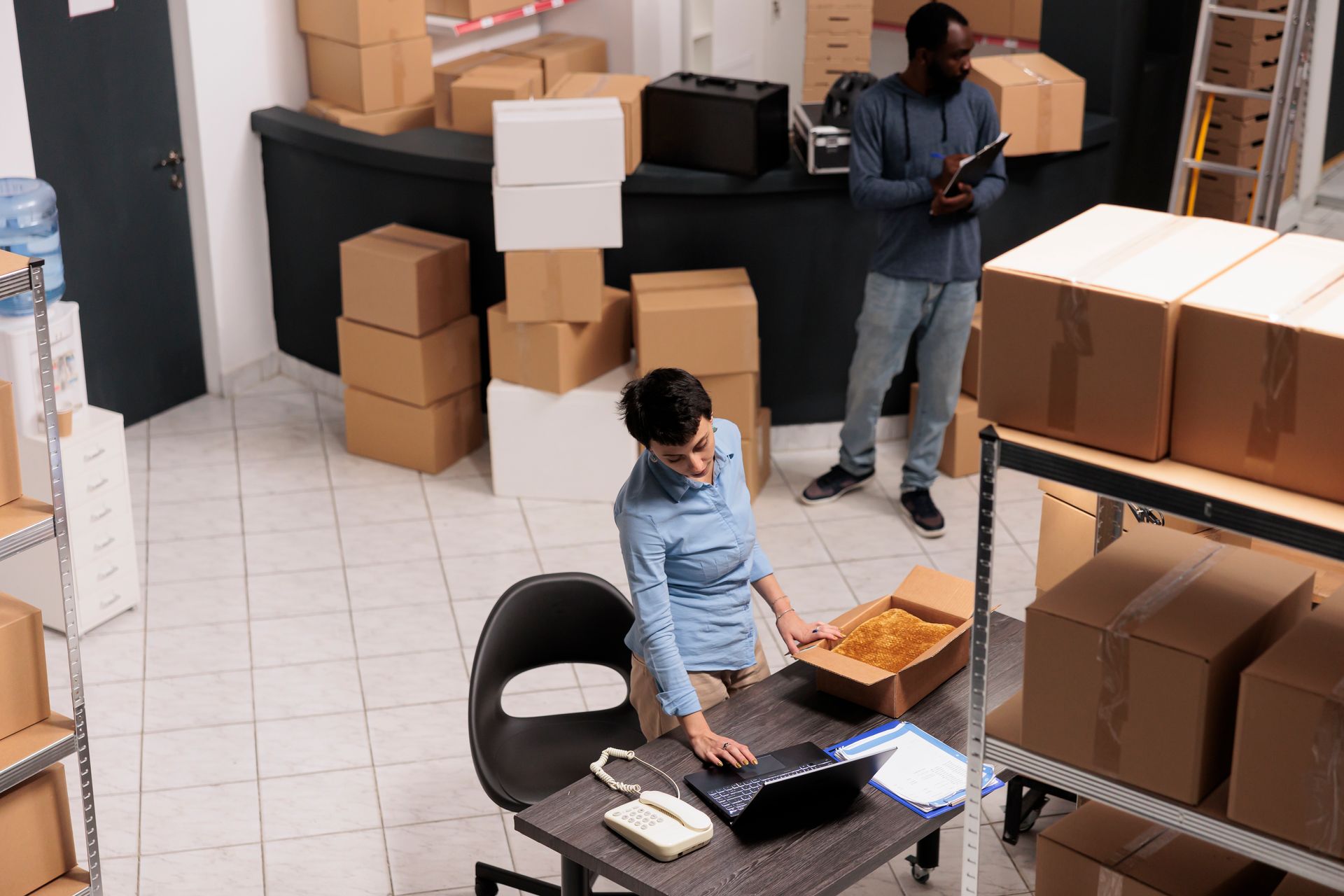 A woman is sitting at a desk in a warehouse using a laptop computer.
