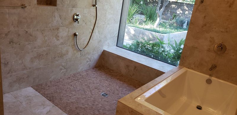 Bathroom shower with beige stone walls, pebble floor, and a large window overlooking greenery. White bathtub visible.