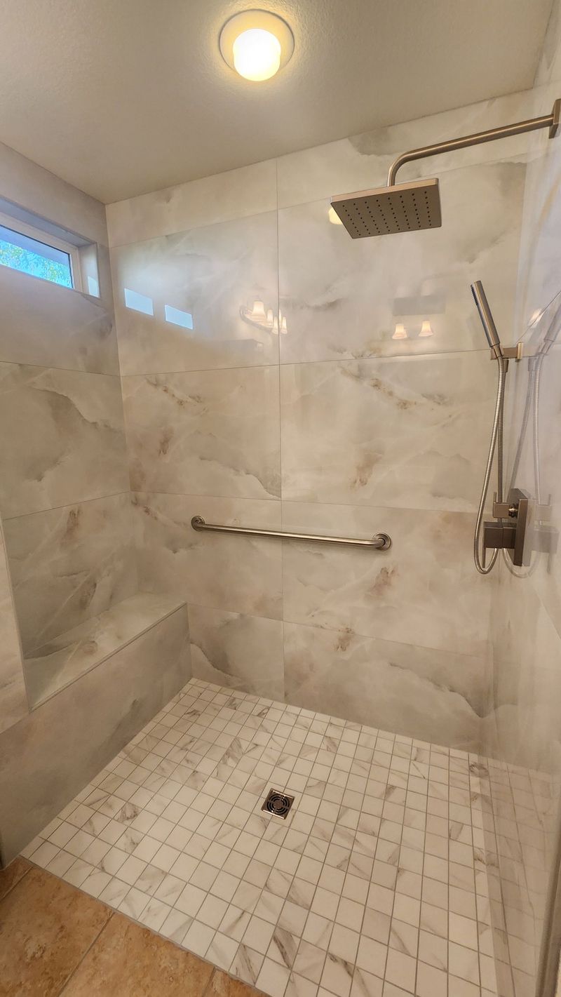 Accessible shower with marble-like walls, mosaic floor, handrail, and rain shower head.