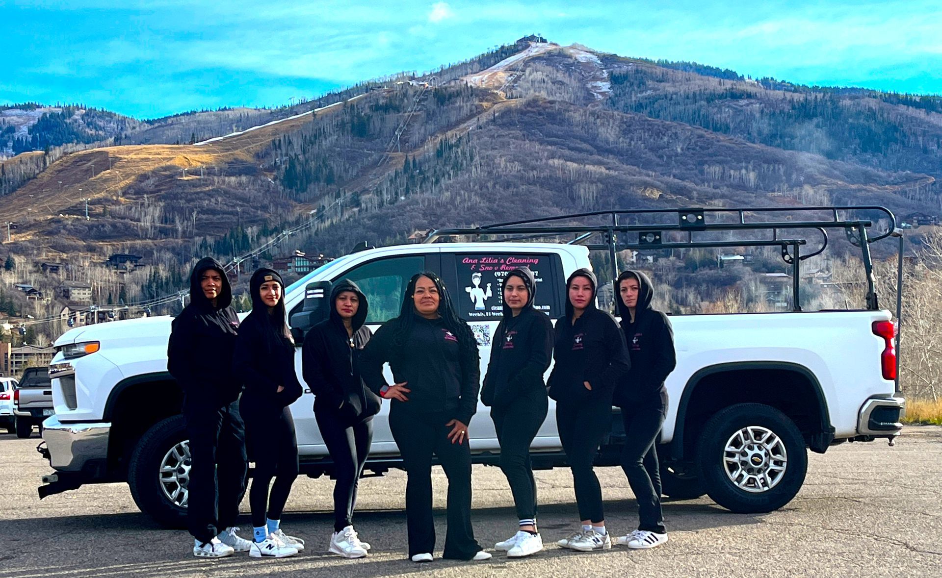 Group of people in black hoodies stand in front of a white truck, mountain in the background.