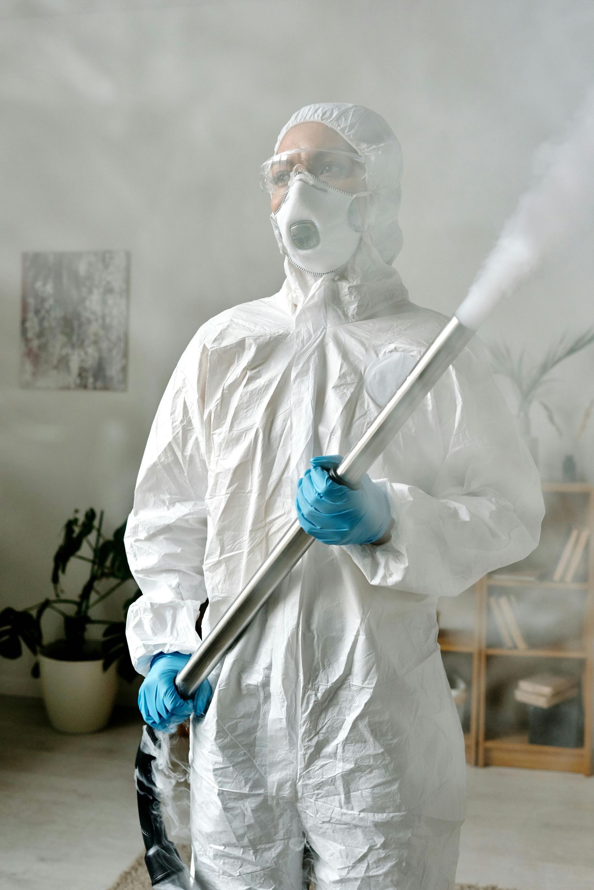 A worker in a protective suit and mask cleaning black mold from a white wall inside a building near a window.