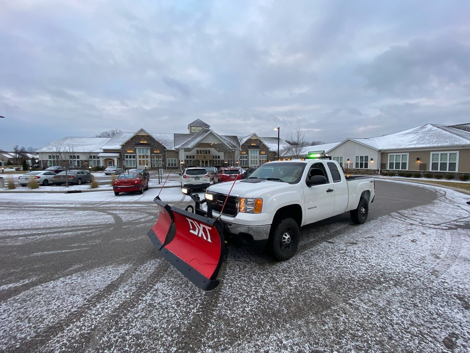 White pickup truck with a red snowplow clearing a snowy parking lot. Buildings in the background.