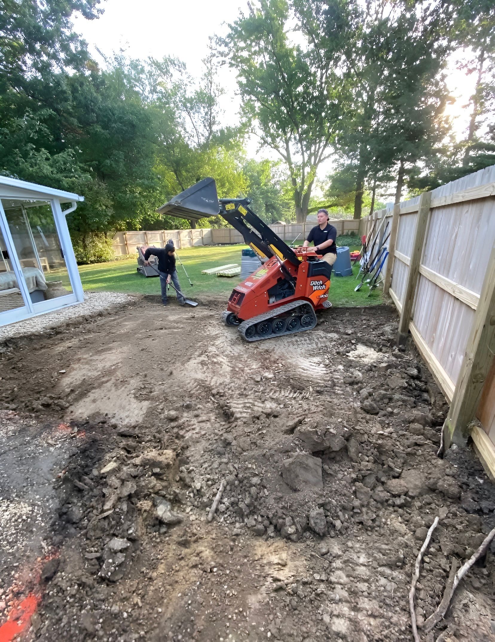 Two people excavating a backyard with a mini excavator and shovels. Soil, trees, and a fence are visible.