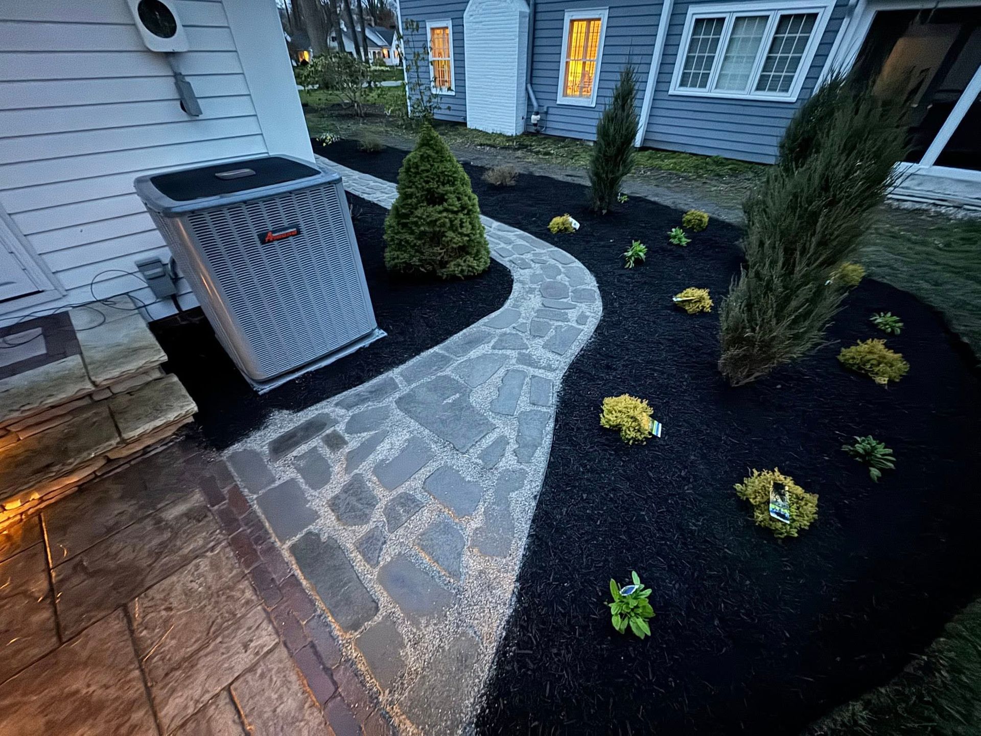 Stone pathway through a garden bed with black mulch, leading to a gray house. An air conditioning unit sits next to the pathway.