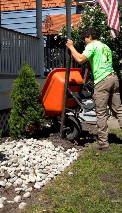Man in green shirt using a wheelbarrow to move material in a garden bed next to a house.