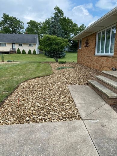 Front yard with stone landscaping next to a concrete walkway leading to a brick house. Green grass and trees are in the background.
