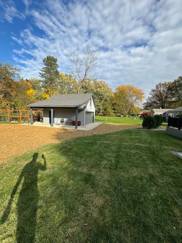 Gray shed with open covered area, on a grassy yard, under a partly cloudy blue sky. Shadow of a person taking the photo.