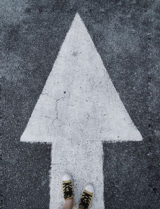 White arrow painted on dark pavement, with the toes of two shoes visible at the bottom.