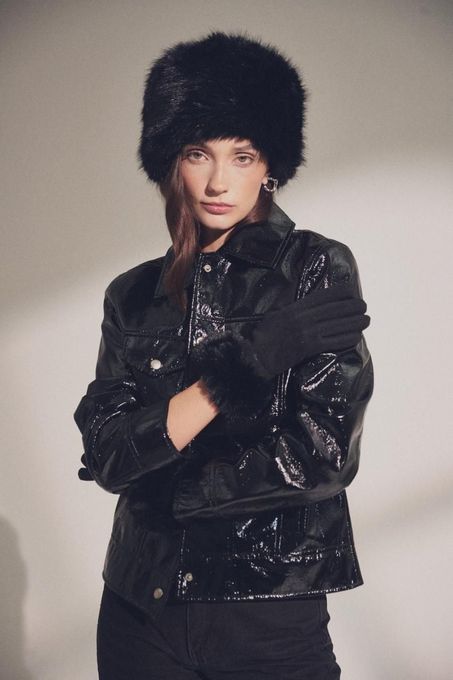 Una persona con un gorro negro de piel sintética y una chaqueta negra texturizada sobre un fondo liso de tonos claros.