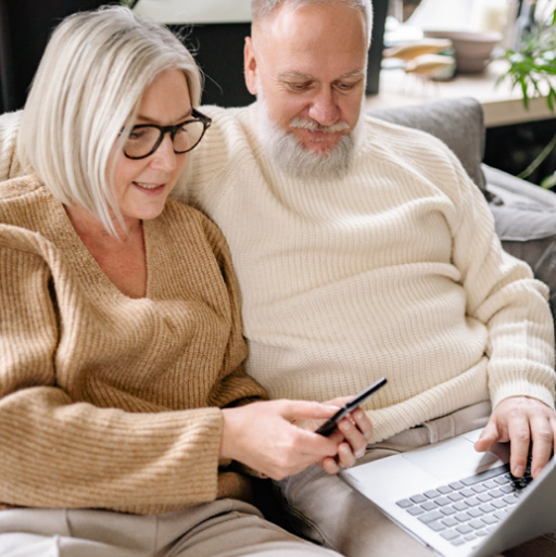 Older couple on couch looking at phone and laptop. Woman holds phone, man uses laptop. Indoors.
