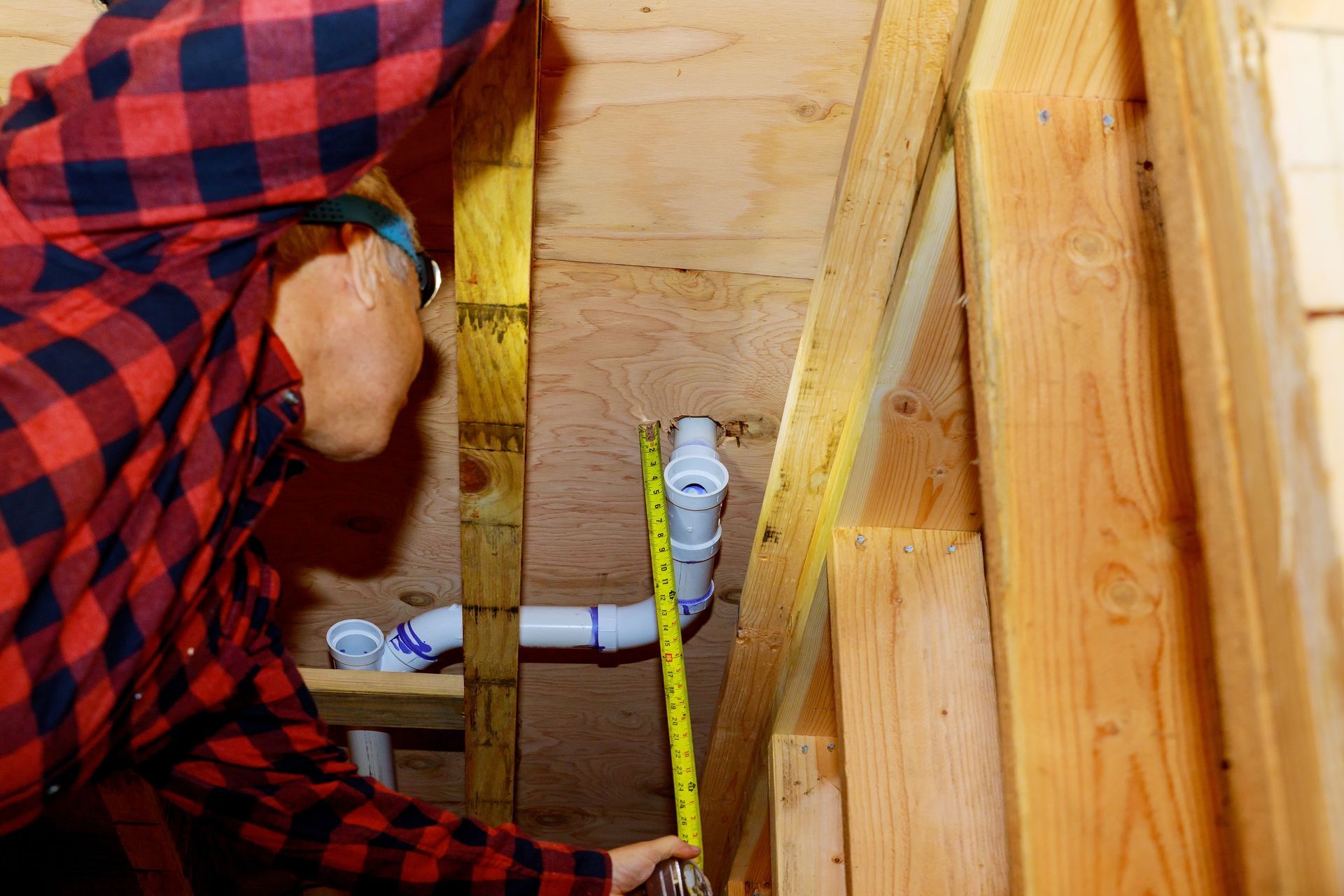 A man in a plaid shirt is measuring a pipe with a tape measure.