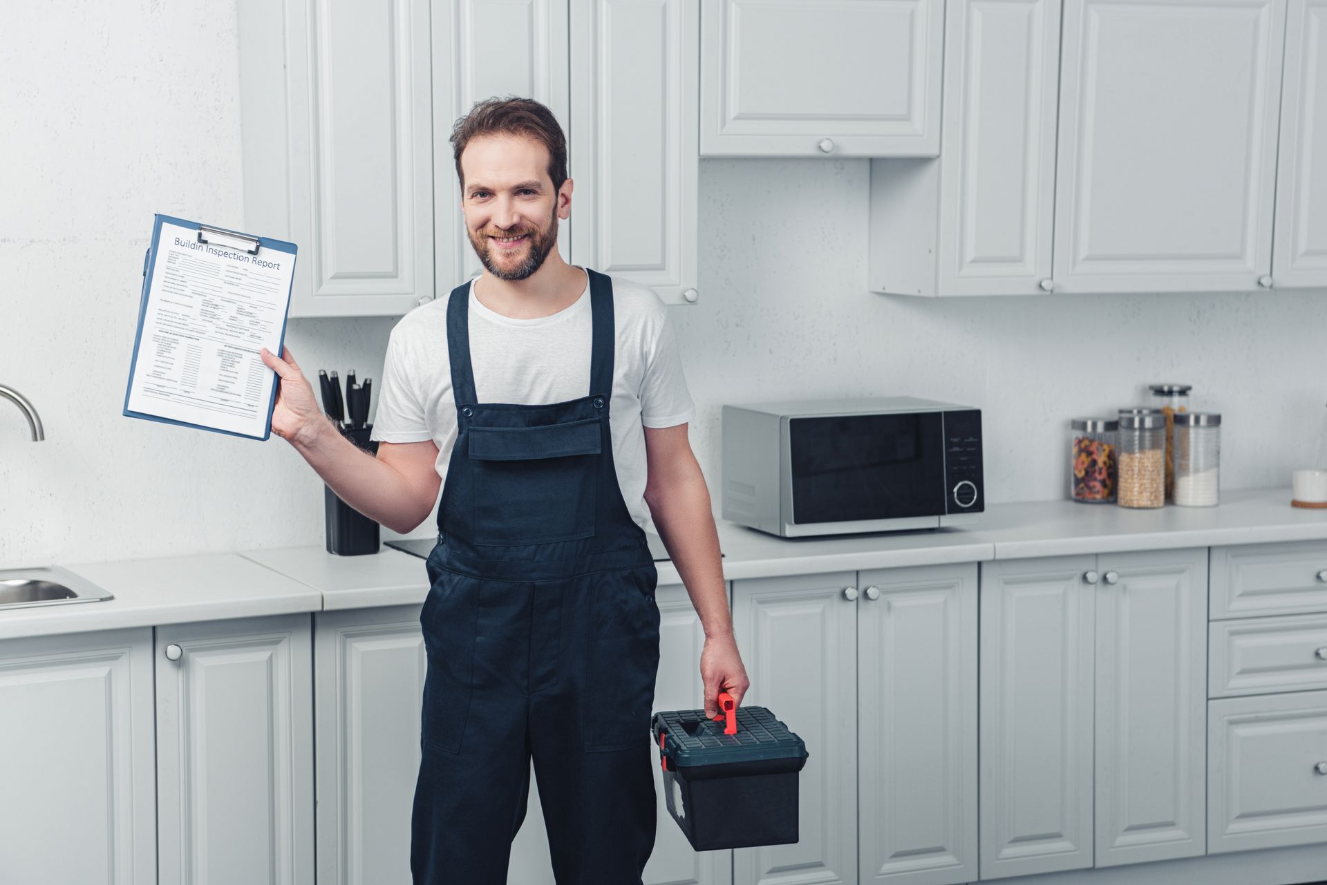 A man in overalls is holding a clipboard and a toolbox in a kitchen.