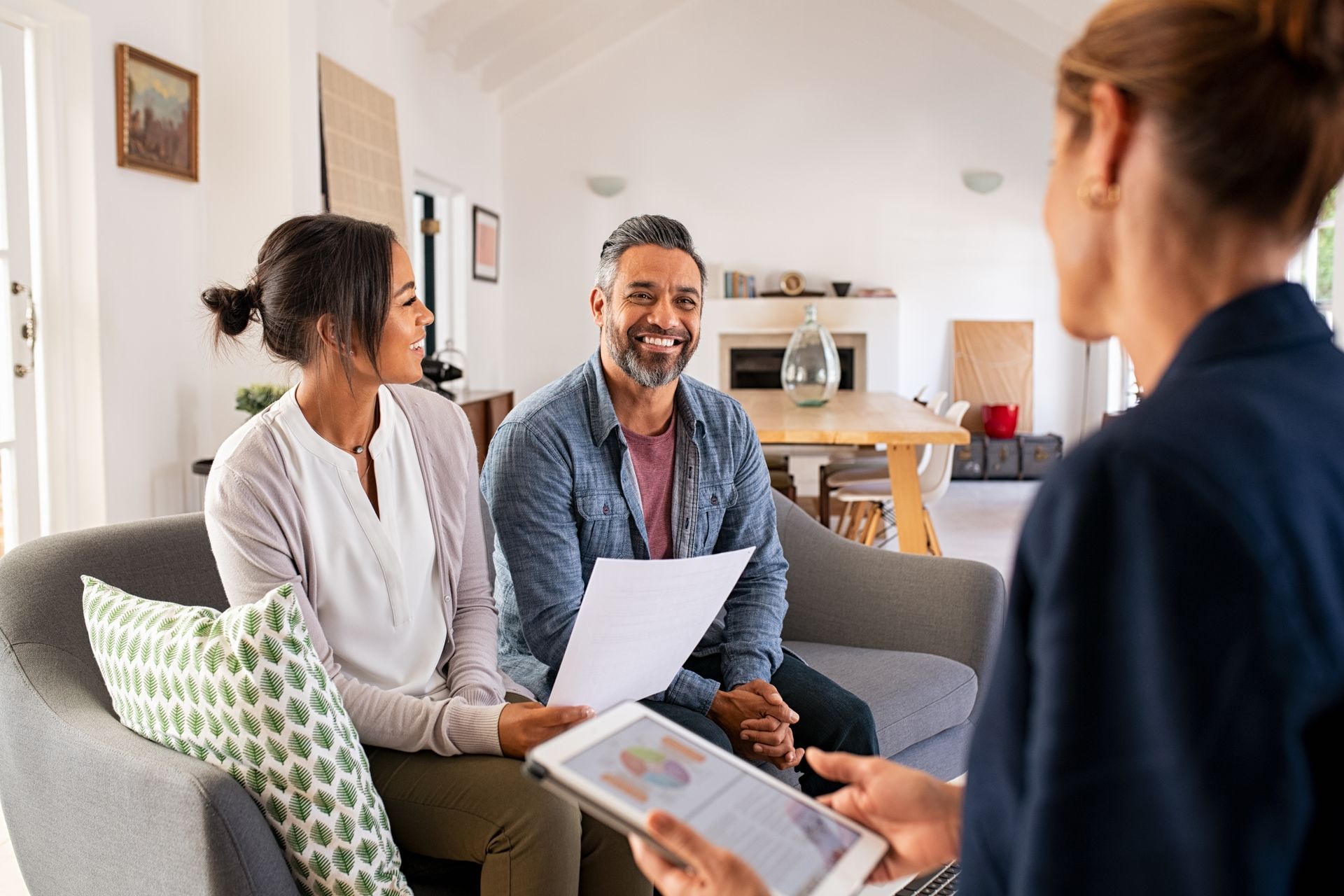 Couple on couch smiling at person holding tablet and papers